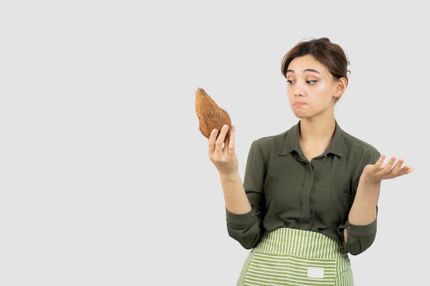 Portrait of young woman in apron holding a coconut against white wall