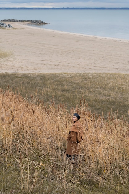 Portrait of young woman in a black shiny beret and a beige wool coat posing against the autumn field