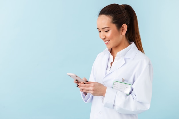 portrait of a young woman doctor posing isolated over blue wall using mobile phone.