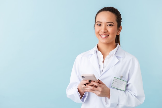 portrait of a young woman doctor posing isolated over blue wall using mobile phone.