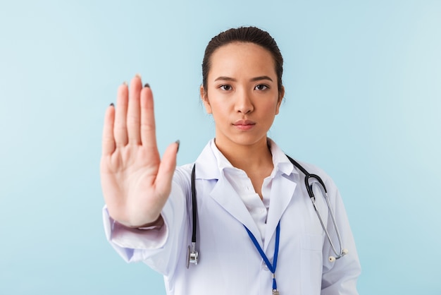 portrait of a young woman doctor posing isolated over blue wall with stethoscope showing stop gesture.