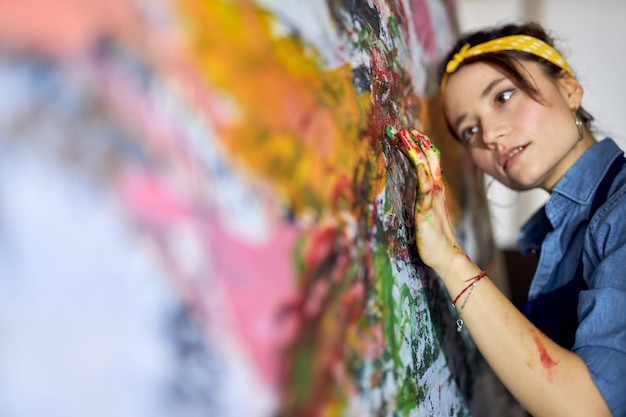 Portrait of young woman female painter applying paint on canvas with fingers while creating a large