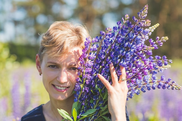 Portrait of young woman holding bouquet of lupin flowers walking in summer meadow Stylish girl is enjoying sunny weather blonde woman with bouquet of flowersLupine field Summer vacation