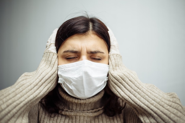 Portrait of a young woman holds hands on her head and has to stay home during quarantine due to coronavirus pandemia. Beautiful girl wearing medical mask and gloves. Covid-19 epidemia concept.