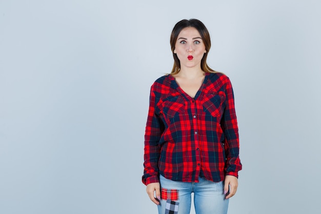 Portrait of young woman keeping lips folded in checked shirt, jeans and looking puzzled front view