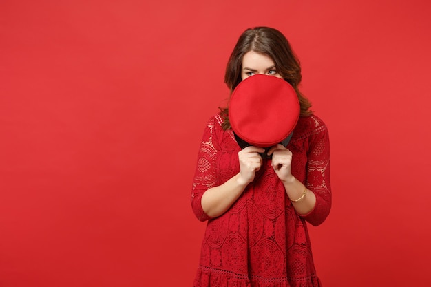 Portrait of young woman in lace dress hiding, covering face with cap, looking aside isolated on bright red wall background in studio. People sincere emotions, lifestyle concept. Mock up copy space.