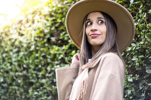 Portrait of a young woman looking at the camera and holding her hat with green leaves in the background outdoors