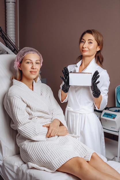 Portrait of a young woman lying in a cross and a master cosmetologist standing next to her holding a copy paste box