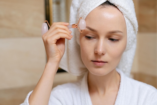 Photo portrait of a young woman massaging her face with a rose quartz gouache roller in the bathroom the concept of cosmetic beauty procedures