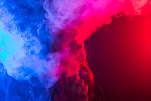 Portrait of young woman in neon red and blue smoke with vape or ecigarettes.