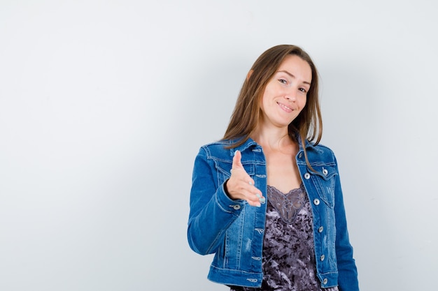 Portrait of young woman offering handshake for greeting in denim jacket, dress and looking gentle front view