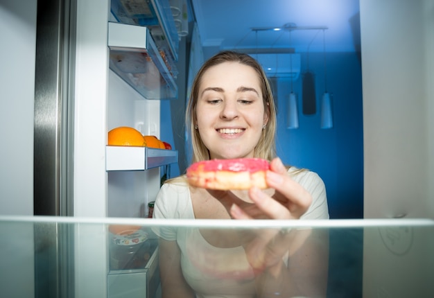 Portrait of young woman opening refrigerator and taking big donut at night