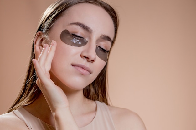 Portrait of young woman posing with applied black beauty patches under eyes over beige background.