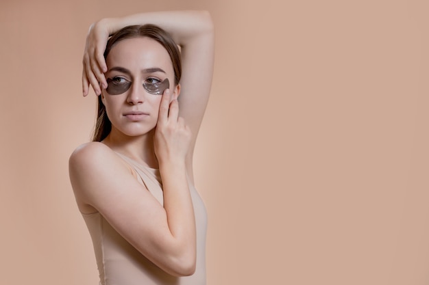 Portrait of young woman posing with applied black beauty patches under eyes over beige background.