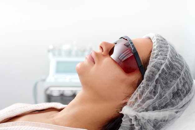 Portrait of young woman in protective eyeglasses lies at couche in beauty salon Concept of professional cosmetology and plastic surgery