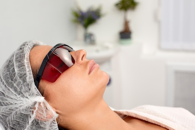 Portrait of young woman in protective eyeglasses lies at couche in beauty salon Concept of professional cosmetology and plastic surgery