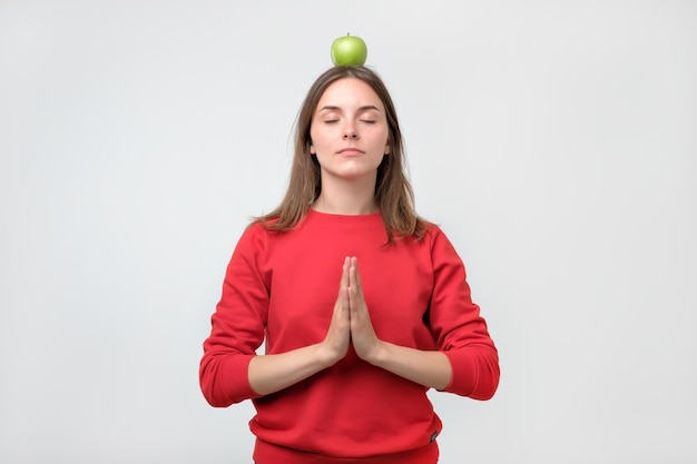 Portrait of young woman in red sweater standing with green apple on head and meditating