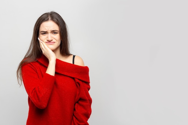 Portrait of a young woman in a red sweater with severe toothache holds her hand on her cheek on a graywhite background