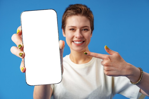 Portrait of a young woman showing blank screen mobile phone while standing over blue background