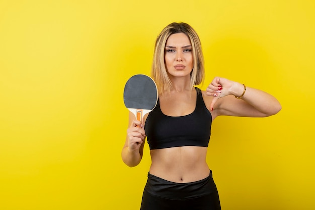 Portrait of young woman showing thumb down and holding table tennis racket.  