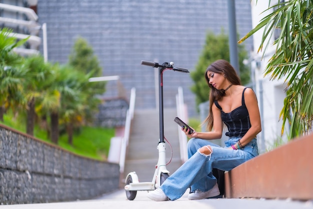 Portrait of a young woman sitting in the city with an electric scooter and typing a message