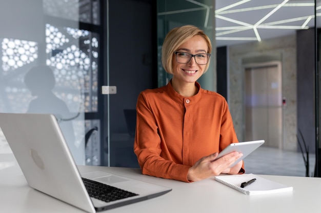 Portrait of a young woman sitting in the office at a table with a laptop and using a tablet he looks