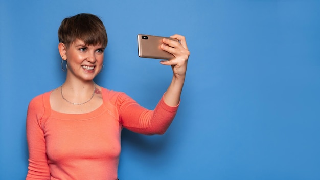 Portrait of a young woman taking a selfie on a smartphone on a blue background. A place for your text. The concept of video communication at a distance.