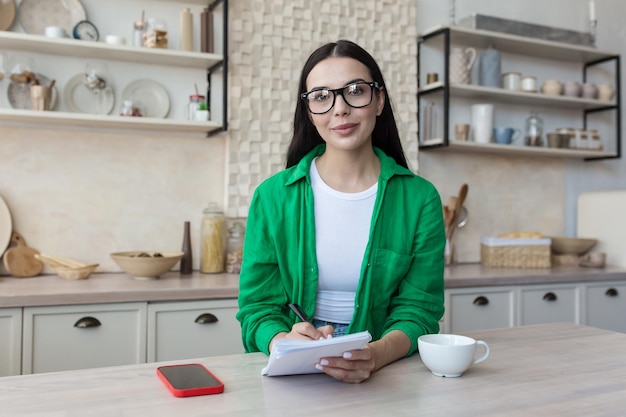 Portrait of a young woman teacher in glasses sitting at home in the kitchen in front of the camera