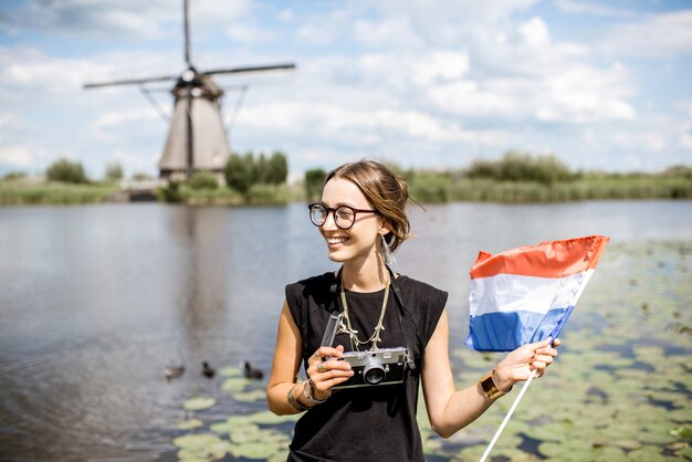 Photo portrait of a young woman tourist standing with dutch flag on the beautiful landscape background with old windmills in netherlands