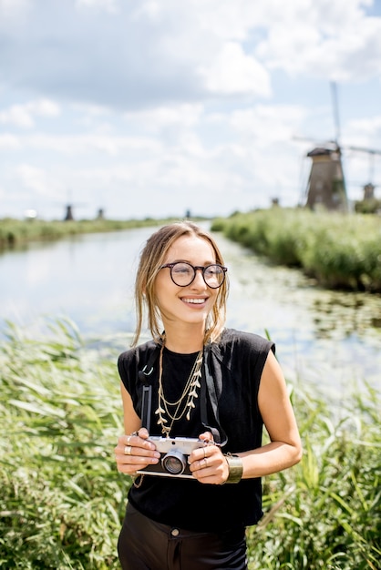 Photo portrait of a young woman tourist standing with photo camera on the beautiful landscape background with old windmills in netherlands