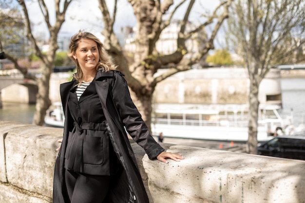 Portrait of young woman wearing black trench coat trousers leaning on concrete fence near river on sunny windy day