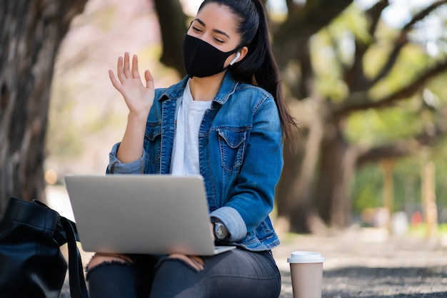 Portrait of young woman wearing face mask on a video call with laptop while sitting outdoors. Urban concept. New normal lifestyle concept.