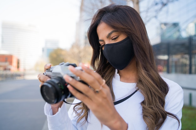 Portrait of young woman wearing protective mask and using camera while taking photographs in the city. New normal lifestyle concept.