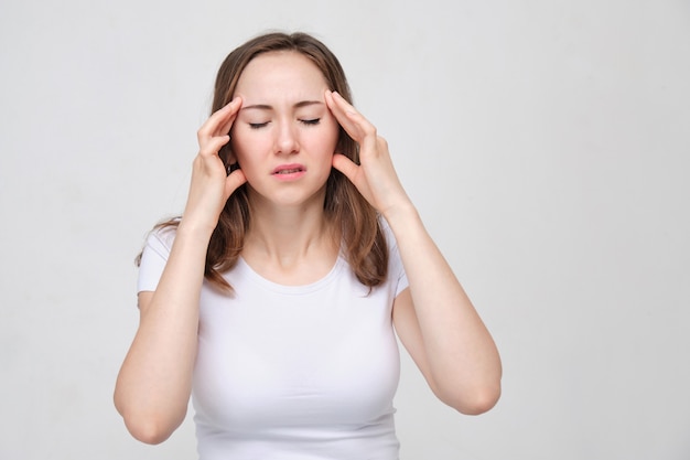 A portrait of a young woman in a white shirt holding his hands on his head. The concept of a strong head pain. headache.