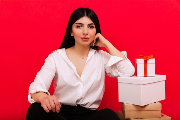 Portrait of a young woman in a white shirt and jeans sitting next to Christmas gifts on a red background