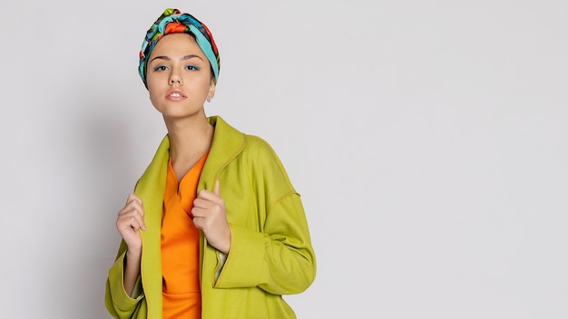 Portrait of a young woman with bright makeup and a fashionable headscarf. Light background. Beauty, fashion, makeup concept. girl in a bright green coat, bright orange dress. 16:9 panoramic format.