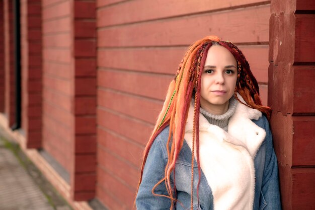 Portrait of young woman with ginger dreadlocks standing leaning on wall on city street Positive female looking at camera smiling