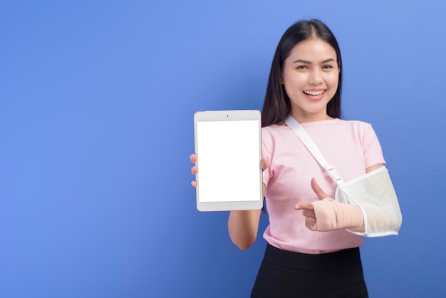 A portrait of young woman with an injured arm in a sling using a tablet over blue background in studio, insurance and healthcare concept