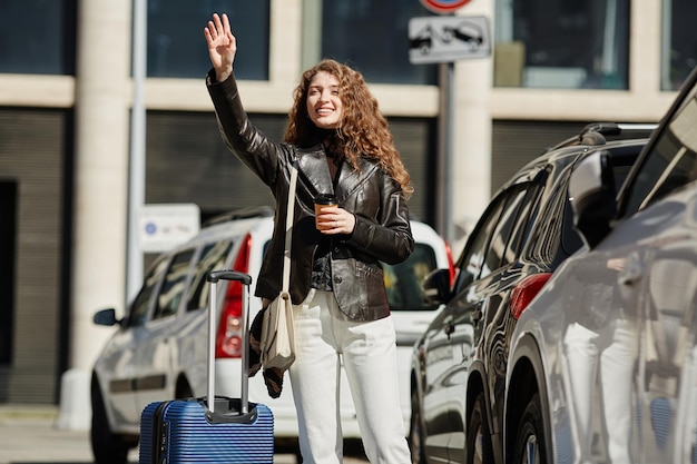 Portrait of young woman with long hair hailing taxi in city street and smiling