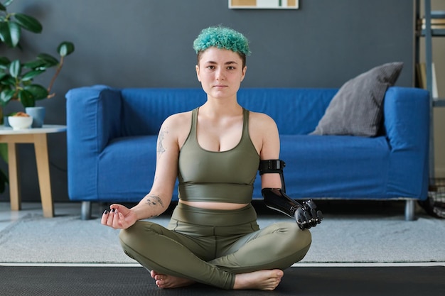 Portrait of young woman with prosthesis sitting on exercise mat in the room and doing yoga