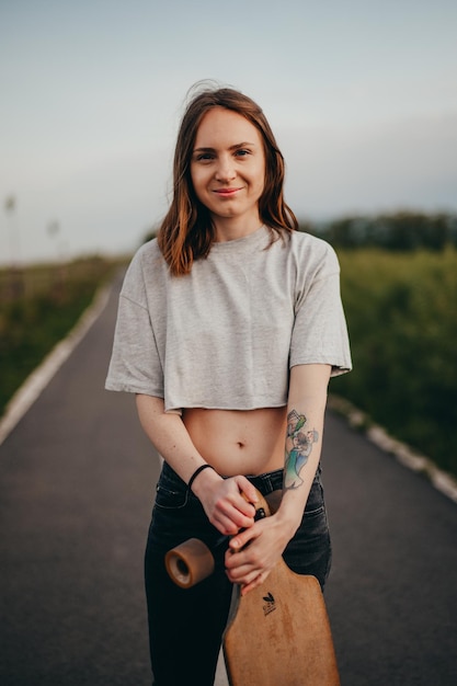 A portrait young woman with tattoo leaning on a longboard looks into the camera