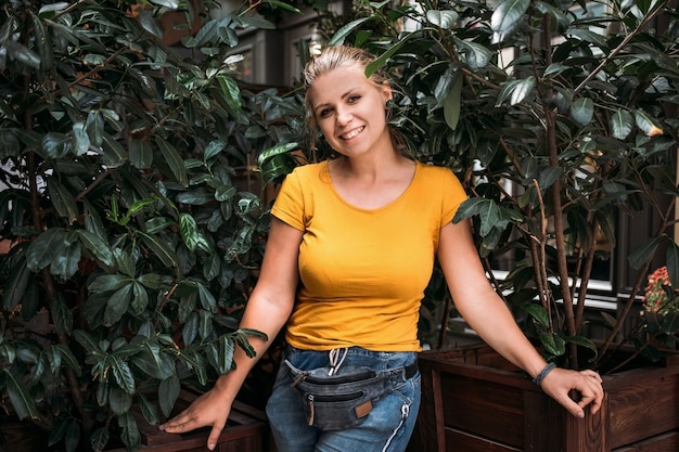 Portrait of a young woman in a yellow T-shirt and blue jeans and a belt bag near flowerpots