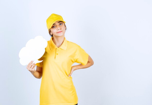 Portrait of a young woman in yellow uniform holding an empty white speech bubble cloud . 