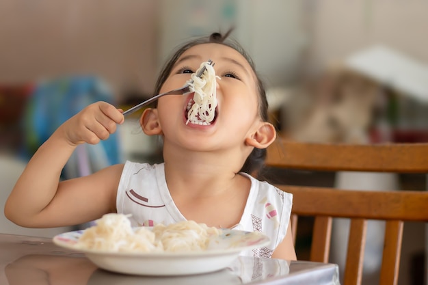 Portraitâ imageâ ofâ 3-4 yearsâ oldâ ofâ baby.â Happyâ Asianâ childâ girlâ enjoyâ eatingâ someâ noodles byâ herself.â Healthyâ Foodâ andâ kidâ concept.