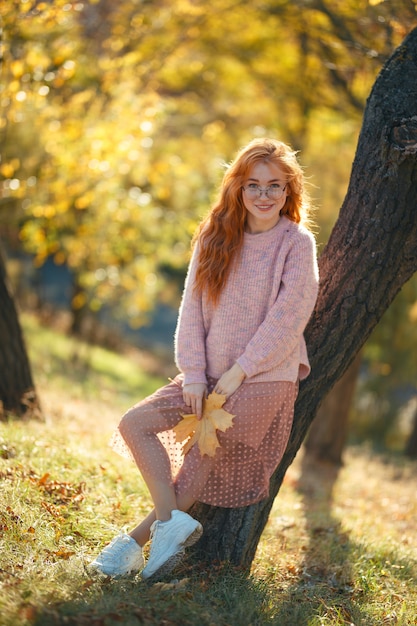 Portraits of a charming red-haired girl with a cute face. Girl posing in autumn park in a sweater and a coral-colored skirt. In the hands of a girl a yellow leaf