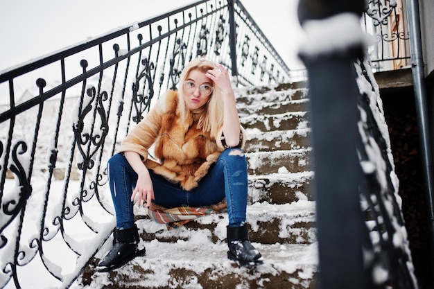 Portraiy of blonde girl in glasses, red fur coat and scarf at winter day.