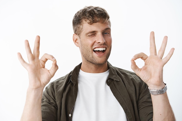 Portrat of good-looking blond man showing okay gesture and winking with broad intriguing smile saying no problem,