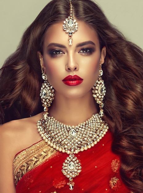 Posh jewelry set is consisting of big earrings, bright necklace and head adornment (tikka). Perfect, dense, wavy, freely flying hair and "smoky eyes" style makeup. Close up portrait.