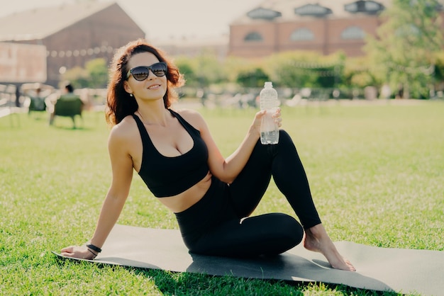 Positive active woman has perfect body shape as result of regular fitness training poses on karemat with bottle of water has rest after workout spends free time outdoors Practicing yoga in morning