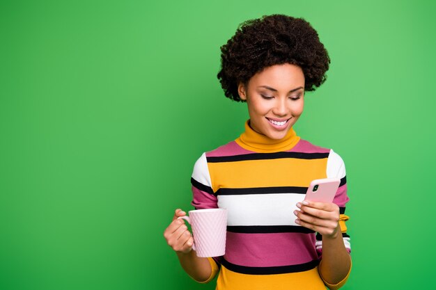 Positive afro american girl use smartphone read social network news follow share comment subscribe blog post relax hold beverage mug wear striped bright outfit 